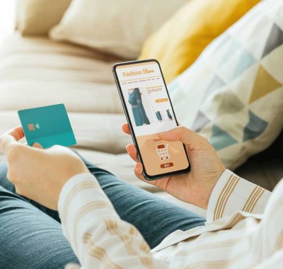 A person is holding a smartphone in one hand and a blue credit card in the other. The phone screen displays an online shopping app with various clothing items and a shopping cart icon. The background features a couch with a patterned pillow.