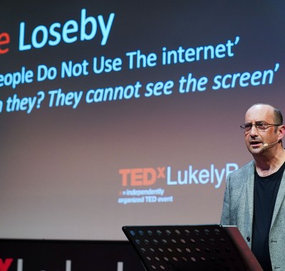 Clive giving his Ted Talk. Behind him, a screen says \Blind People do not use the Internet. How can they? They cannot see the screen\