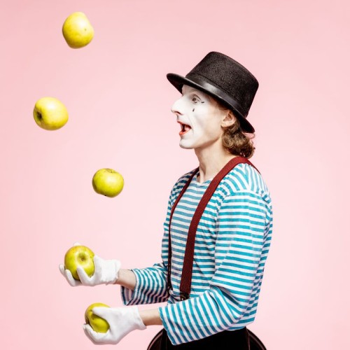 A  clown in a black and white stripy jumper and wearing a hat is juggling apples