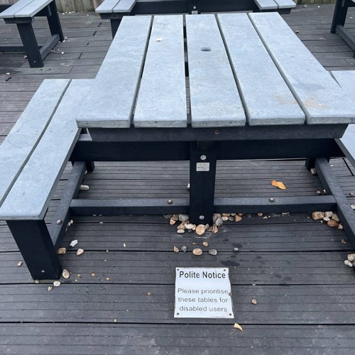 A table with 2 benches fixed to it, gaps are on the other sides for wheelchairs. A sign on the floor reads: Polite Notice Please prioritise these tables for disabled users