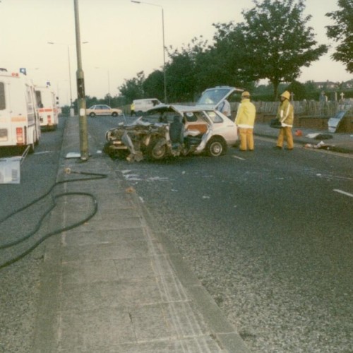 A traffic accident scene showing a significantly damaged car in the middle of the road. Firefighters in yellow gear are on-site, with emergency vehicles including fire engines and an ambulance present. Hoses are laid out on the ground, and trees line the street.