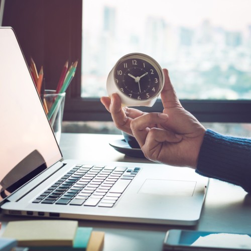 A hand holding a small clock is extended over a laptop on a desk. The desk also has colorful sticky notes, pencils, and a blurry cityscape is visible through the window in the background.