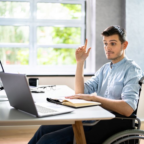 A man sitting in a wheelchair at a desk with a laptop, raising his right hand. He appears to be asking a question or participating in an online meeting. An open notebook and eyeglasses are on the desk. The background shows large windows with greenery outside.