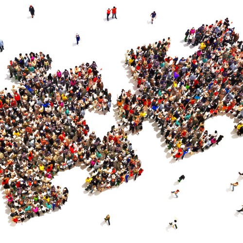 A large group of people is gathered to form two interlocking puzzle pieces against a white background. Additional smaller groups and individuals are scattered around the central formation, creating a sense of movement and interaction among the crowd.