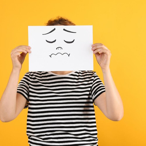 A person wearing a black and white striped shirt holds a white piece of paper with a sad face drawn on it, covering their own face. The background is solid yellow. src=https://accessbydesign.uk/abd/wp-content/gallery/blog-images/Woman-drawing-sad-face-w.jpg