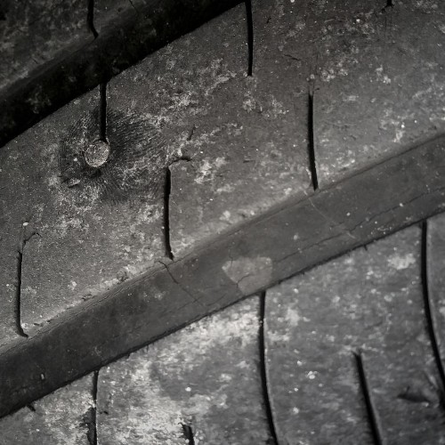A close-up image of a worn car tire with visible cracks and a nail embedded in the tread. The tire\'s surface shows signs of wear and tear, indicating it has been used extensively and requires repair or replacement.