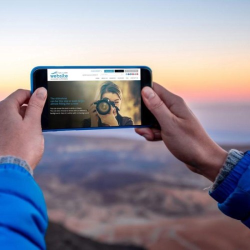 A person wearing a blue jacket holds a smartphone horizontally to take a photo of a scenic landscape at sunrise or sunset. The image on the phone's screen shows another person photographing the viewer. The background features rolling hills under a colorful sky.