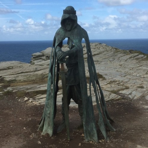 A bronze statue of a cloaked figure stands on a rocky cliffside with the ocean in the background. The cloak appears tattered and the figure holds a staff. The statue is set against a backdrop of a partly cloudy sky and rugged terrain.