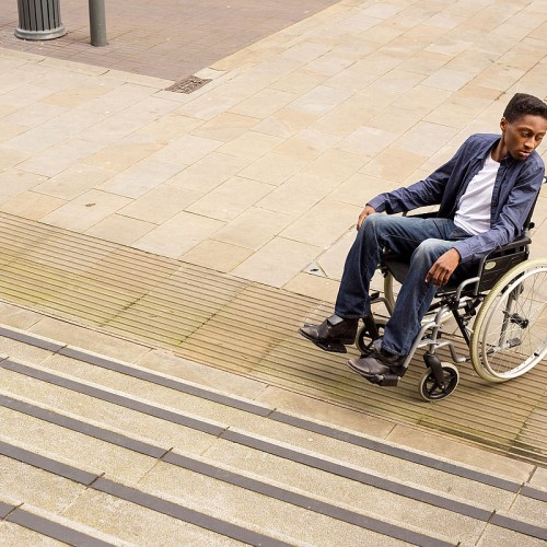 A person in a wheelchair, dressed in a blue blazer and jeans, looks to their right at the base of a flight of stairs. They are on a paved outdoor area with a lamppost in the background.