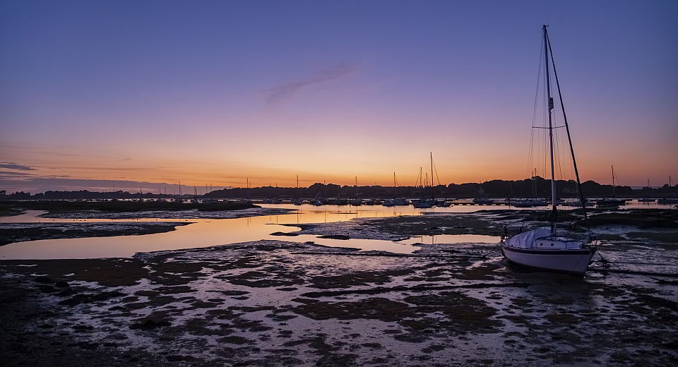 Sailboats rest on wet sand at low tide with their reflections visible in calm water under a purple and orange sunset sky.