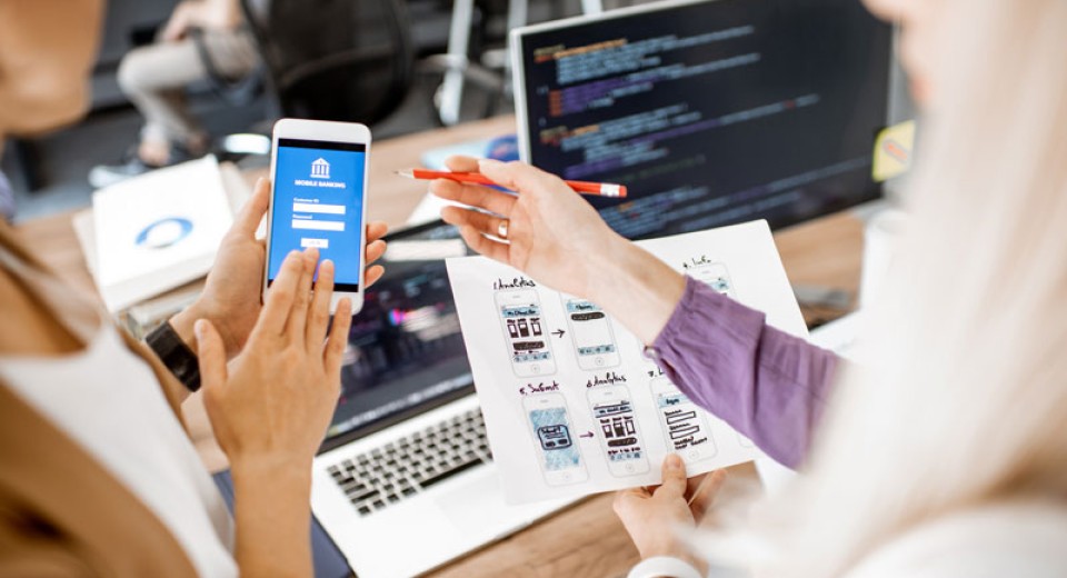 Two people collaborating at a desk with a laptop displaying code, a smartphone showing a banking app screen, and a printed sheet with mobile app wireframes. They are likely discussing a mobile app design and development.