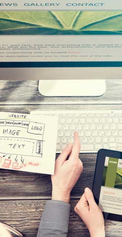 A person is holding a sketch of a website layout above a keyboard on a wooden desk. To the right, part of a tablet screen displays a webpage. Surrounding items include a pencil holder with colorful pencils and a pair of glasses.