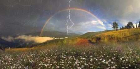 A rainbow in front of dark grey clouds, a lightning bolt is in front of it