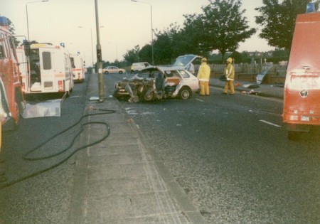 A traffic accident scene showing a significantly damaged car in the middle of the road. Firefighters in yellow gear are on-site, with emergency vehicles including fire engines and an ambulance present. Hoses are laid out on the ground, and trees line the street.