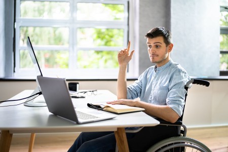 A man sitting in a wheelchair at a desk with a laptop, raising his right hand. He appears to be asking a question or participating in an online meeting. An open notebook and eyeglasses are on the desk. The background shows large windows with greenery outside.