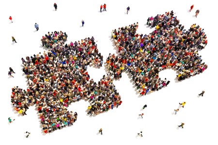 A large group of people is gathered to form two interlocking puzzle pieces against a white background. Additional smaller groups and individuals are scattered around the central formation, creating a sense of movement and interaction among the crowd.