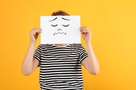 A person wearing a black and white striped shirt holds a white piece of paper with a sad face drawn on it, covering their own face. The background is solid yellow. src=https://accessbydesign.uk/abd/wp-content/gallery/blog-images/Woman-drawing-sad-face-w.jpg