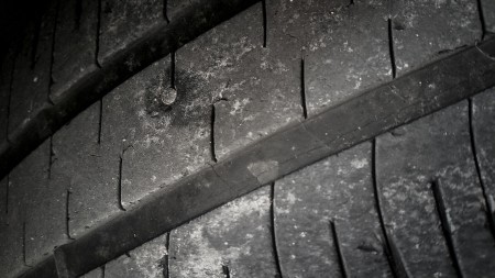 A close-up image of a worn car tire with visible cracks and a nail embedded in the tread. The tire's surface shows signs of wear and tear, indicating it has been used extensively and requires repair or replacement.