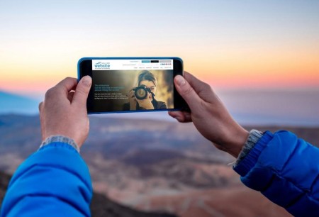 A person wearing a blue jacket holds a smartphone horizontally to take a photo of a scenic landscape at sunrise or sunset. The image on the phone's screen shows another person photographing the viewer. The background features rolling hills under a colorful sky.