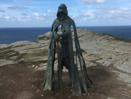 A bronze statue of a cloaked figure stands on a rocky cliffside with the ocean in the background. The cloak appears tattered and the figure holds a staff. The statue is set against a backdrop of a partly cloudy sky and rugged terrain.