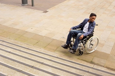 A person in a wheelchair, dressed in a blue blazer and jeans, looks to their right at the base of a flight of stairs. They are on a paved outdoor area with a lamppost in the background.