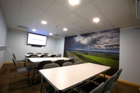 A conference room with white rectangular tables and gray chairs in front of a mural depicting a landscape with green fields and a cloudy sky. There is a wall-mounted screen displaying the text ONE O FOUR. The room has a modern ceiling with recessed lighting.