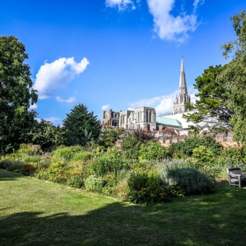 Chichester Cathedral. In the foreground is a lawn and some trees, under a blue sky
