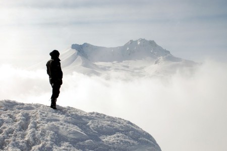 Man on a mountain is gazing into the distance, across a misty landscape. A snow-covered mountain range is far off.