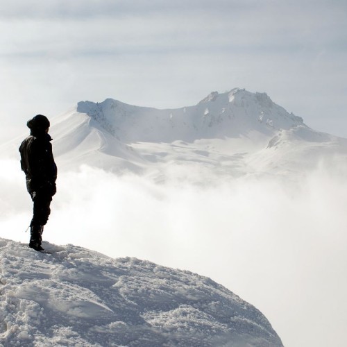 Man on a mountain is gazing into the distance, across a misty landscape. A snow-covered mountain range is far off.