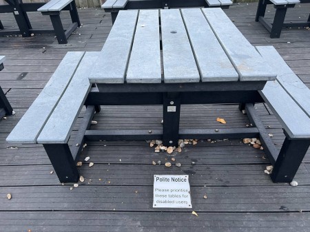 A table with 2 benches fixed to it, gaps are on the other sides for wheelchairs. A sign on the floor reads “Polite Notice Please prioritise these tables for disabled users”
