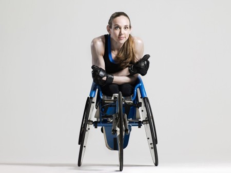 a young female athlete is sitting in a racing wheelchair. She has long hair, a pony tail, blue eyes, no legs and is wearing black leather gloves.