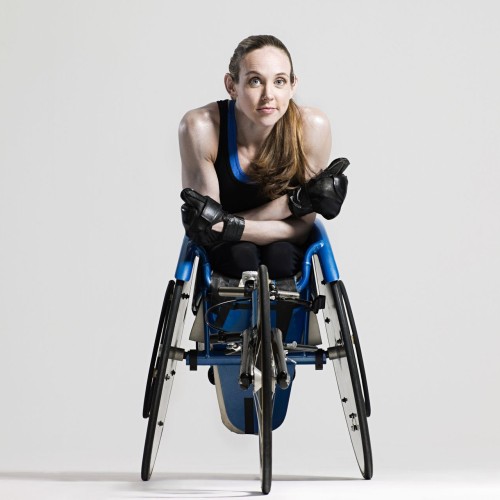a young female athlete is sitting in a racing wheelchair. She has long hair, a pony tail, blue eyes, no legs and is wearing black leather gloves.