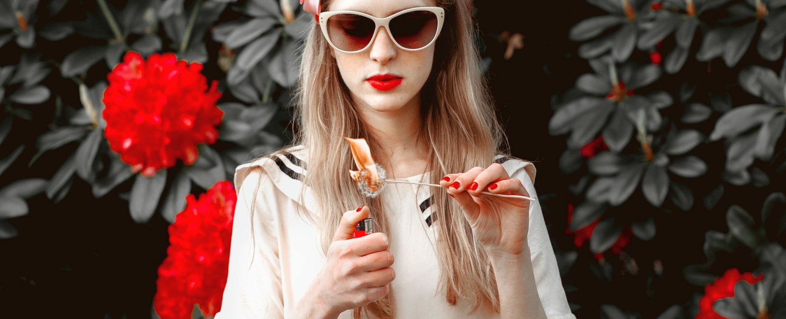 A young woman wearing sunglasses setting a dandelion alight with a cigarette lighter