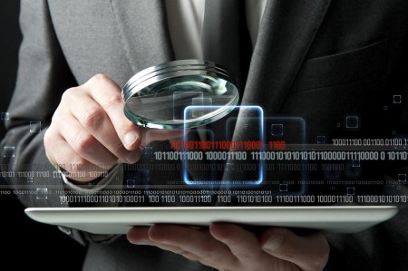 Close-up of a man in a suit holding a magnifying glass over a computer tablet with binary digits overlaid across the image