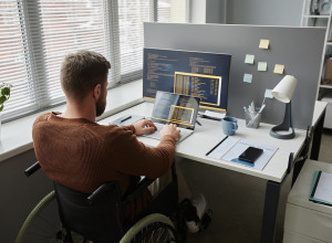 Back view of adult man with disability using wheelchair at office workplace and coding in IT development