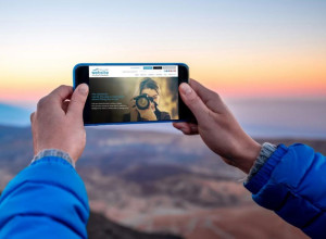 A person wearing a blue jacket holds a smartphone horizontally to take a photo of a scenic landscape at sunrise or sunset. The image on the phone's screen shows another person photographing the viewer. The background features rolling hills under a colorful sky.