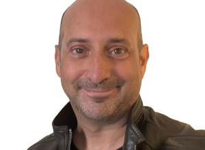 Clive has brown eyes, no hair, has shaped stubble and is smiling