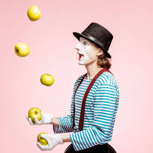 A  clown in a black and white stripy jumper and wearing a hat is juggling apples