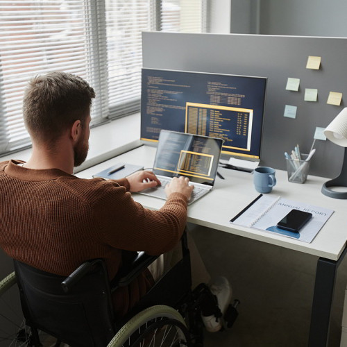 Back view of adult man with disability using wheelchair at office workplace and coding in IT development