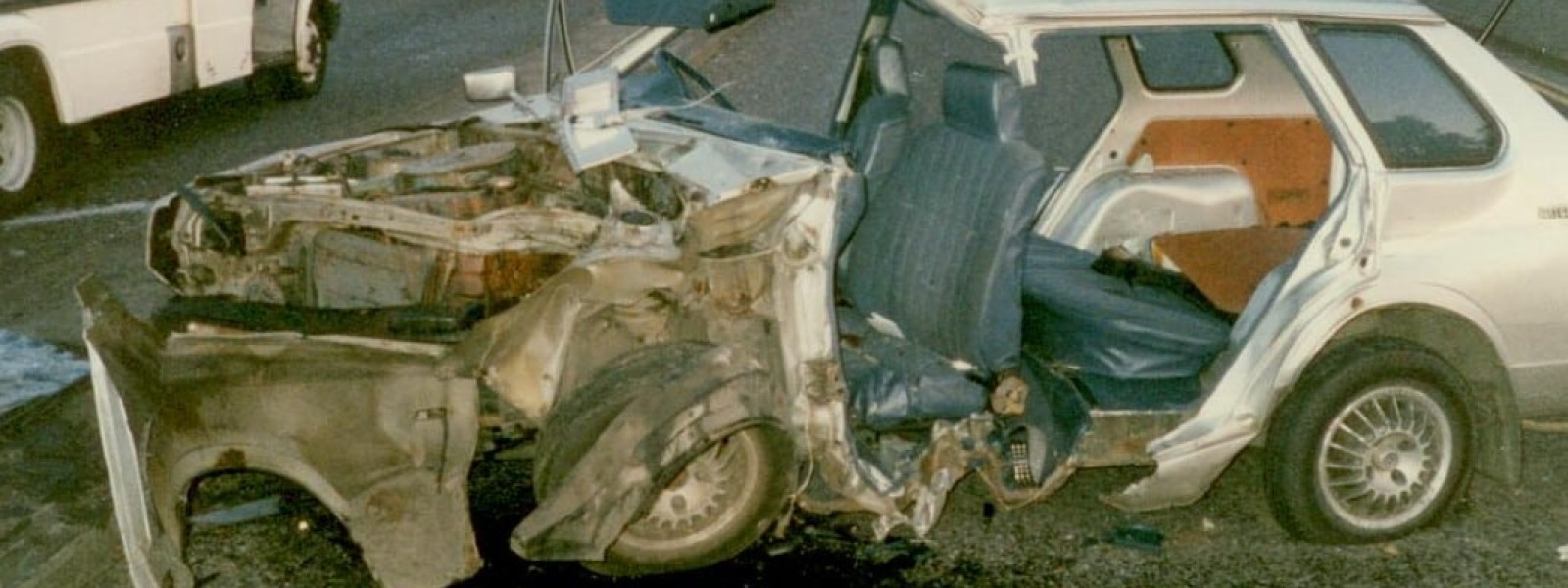 A heavily damaged silver car is shown after a severe collision. The front of the vehicle is extensively wrecked, with parts of the engine compartment exposed. An ambulance and rescue personnel are visible in the background.