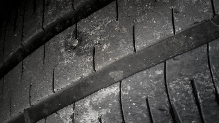 A close-up image of a worn car tire with visible cracks and a nail embedded in the tread. The tire\'s surface shows signs of wear and tear, indicating it has been used extensively and requires repair or replacement.