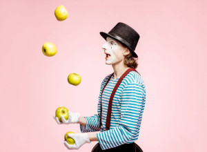 A  clown in a black and white stripy jumper and wearing a hat is juggling apples