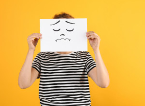 A person wearing a black and white striped shirt holds a white piece of paper with a sad face drawn on it, covering their own face. The background is solid yellow. data-src=https://accessbydesign.uk/abd/wp-content/gallery/blog-images/Woman-drawing-sad-face-w.jpg