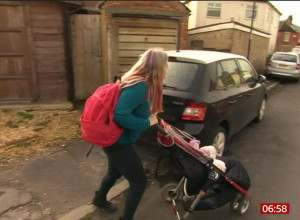 A person with long hair, wearing a turquoise sweater and a red backpack, is pushing a baby stroller down a residential street. The image is timestamped 06:58 in the bottom right corner. Various parked cars and houses are visible in the background.