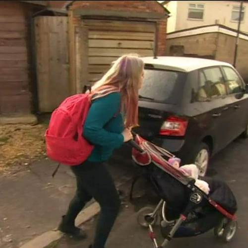 A person with long hair, wearing a turquoise sweater and a red backpack, is pushing a baby stroller down a residential street. The image is timestamped 06:58 in the bottom right corner. Various parked cars and houses are visible in the background.